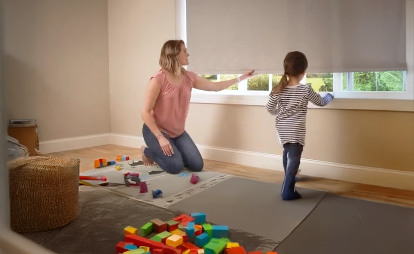 Cordless roller shades like this one enhance the safety of kids' rooms. Add motorization for even more convenience. Mother adjusting window shade while her child plays.